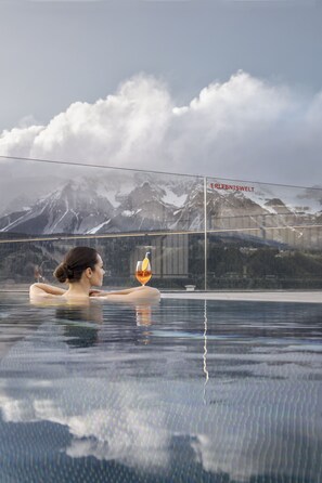 A person relaxing in a glass-bottomed infinity pool with a mountain view and a glass wall displaying the word "ERLEBNISWELT". - Hotel Erlebniswelt Stocker (Schladming)