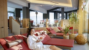 Una mujer con bata blanca sentada en una silla reclinable roja en un espacio interior moderno con detalles de madera y ventanas amplias.