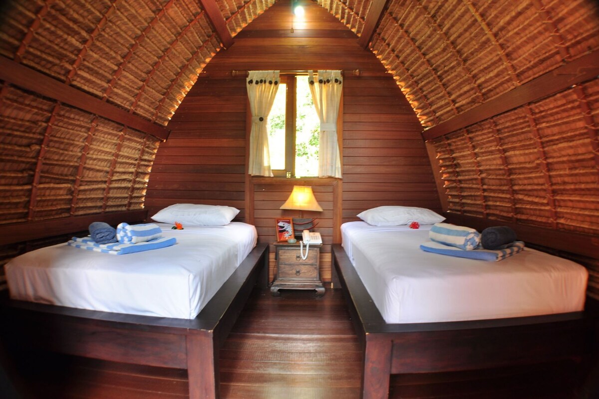A room with two beds, wooden walls, a thatched roof, and a nightstand with a lamp.