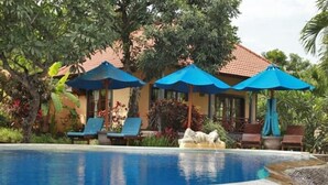 Una piscina con sillones de descanso y sombrillas, rodeada de vegetación tropical y un edificio con tejado de baldosas.