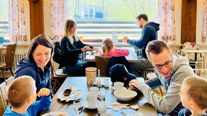 A family dining at a table with floral curtains and a view of a house and trees outside. - Tanneneck (Löffingen)