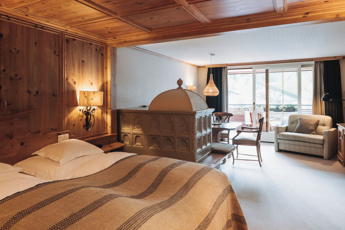 Une chambre d’hôtel avec un grand lit, une tête de lit en bois, un bureau avec une lampe, une chaise et une vue sur l’extérieur grâce à de grandes fenêtres.