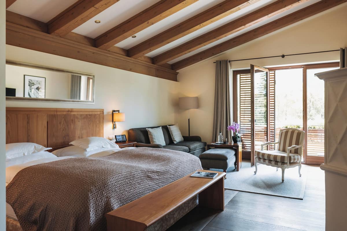 Une chambre avec un grand lit, une tête de lit en bois, un banc, une table de chevet, une lampe, un canapé, un fauteuil et une vue sur l’extérieur par une fenêtre dotée de volets en bois.