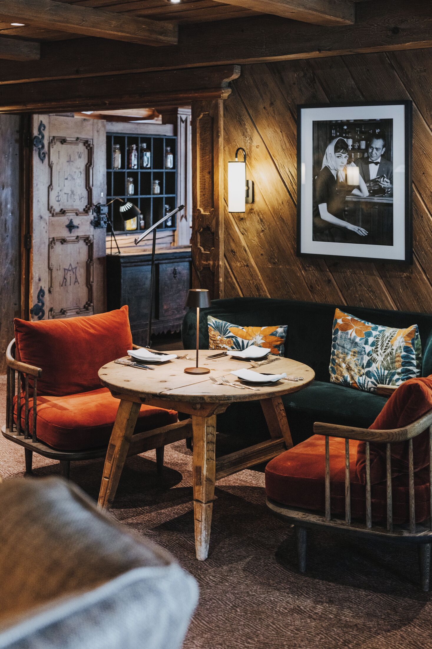 Een knus vertrek met een houten plafond, een ronde houten tafel met stoelen, een bank met kleurrijke kussens en een lijst met een afbeelding aan de muur.