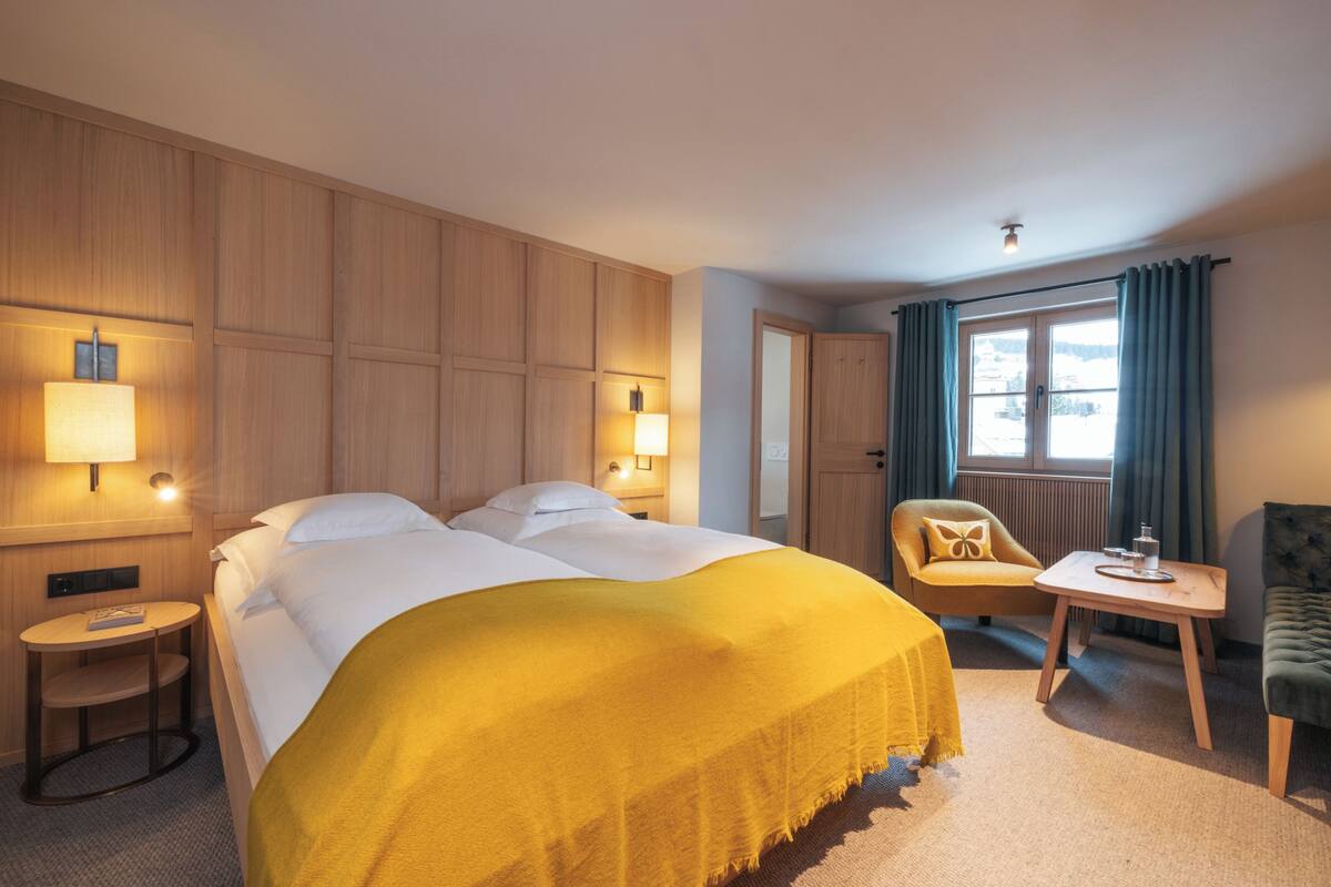 Une chambre d’hôtel avec un grand lit, deux lampes de chevet, une petite table ronde, une chaise et une vue sur l’extérieur par une fenêtre.