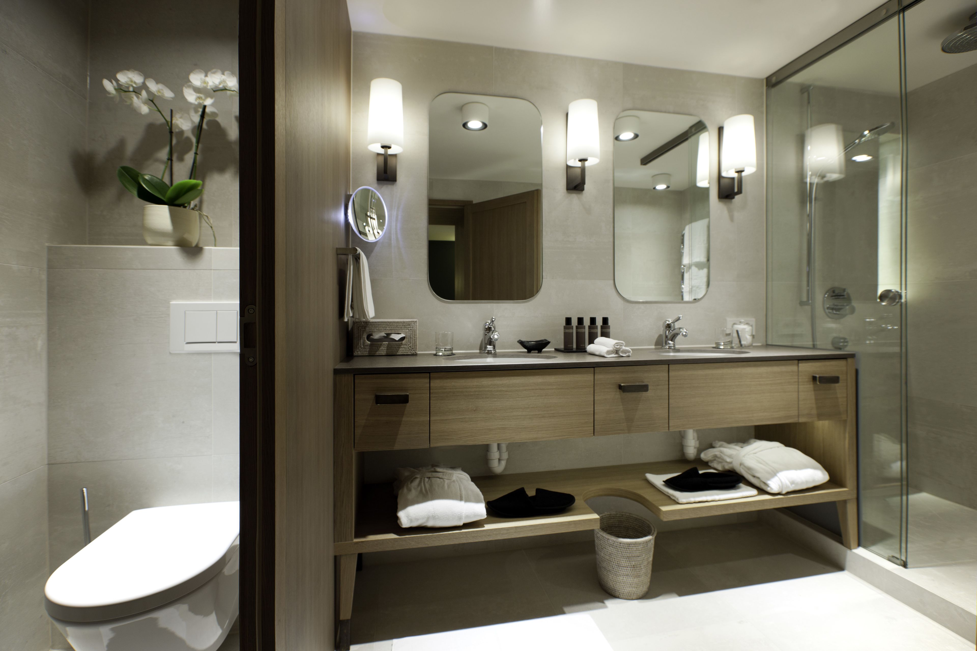 A modern bathroom with a toilet, sink, and shower. It features a vanity with a mirror, shelves, and a potted plant.