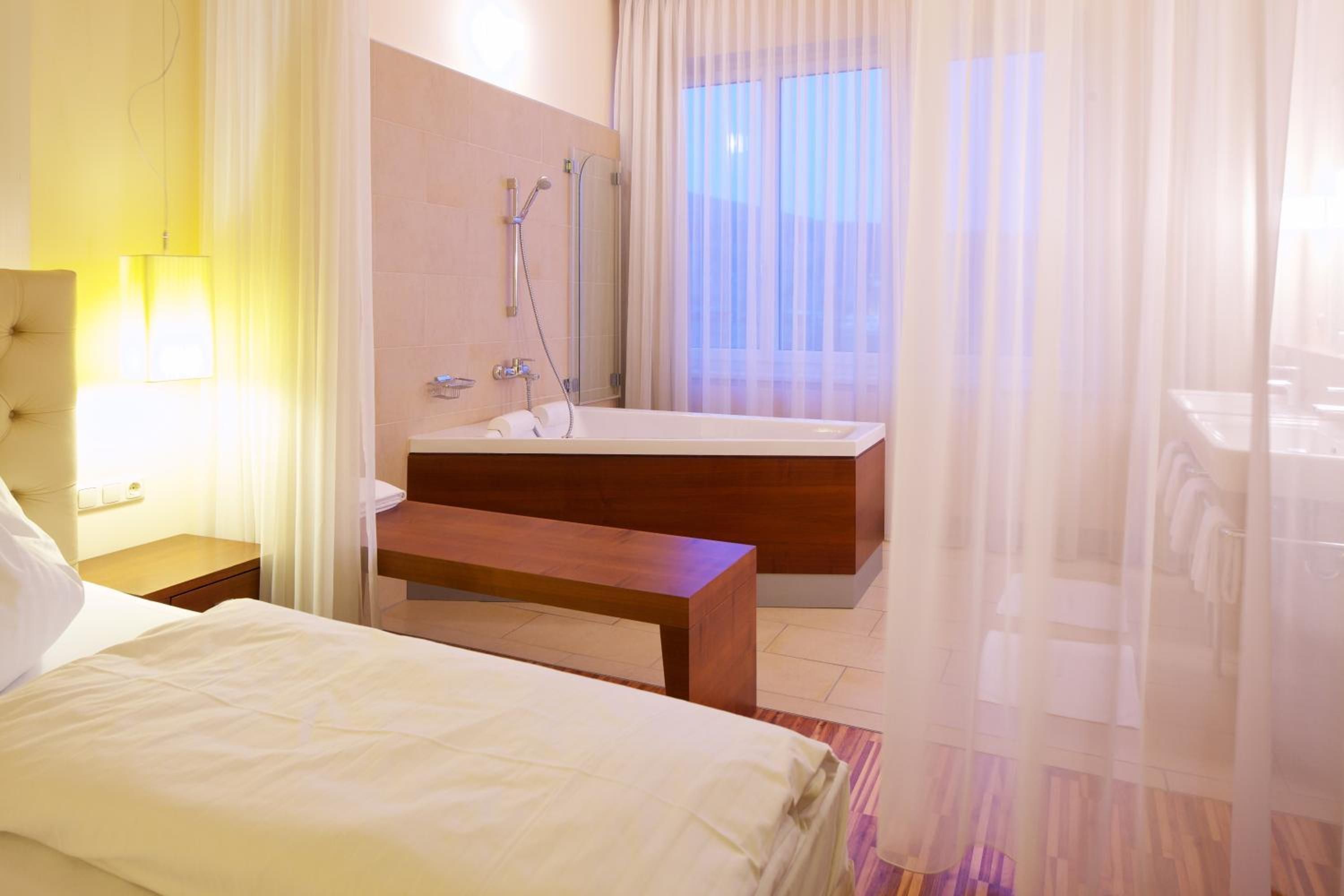 A hotel room with a bed, a wooden bench, a bathroom with a bathtub, and a large window with sheer curtains.