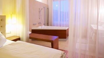 A hotel room with a bed, a wooden bench, a bathroom with a bathtub, and a large window with sheer curtains.