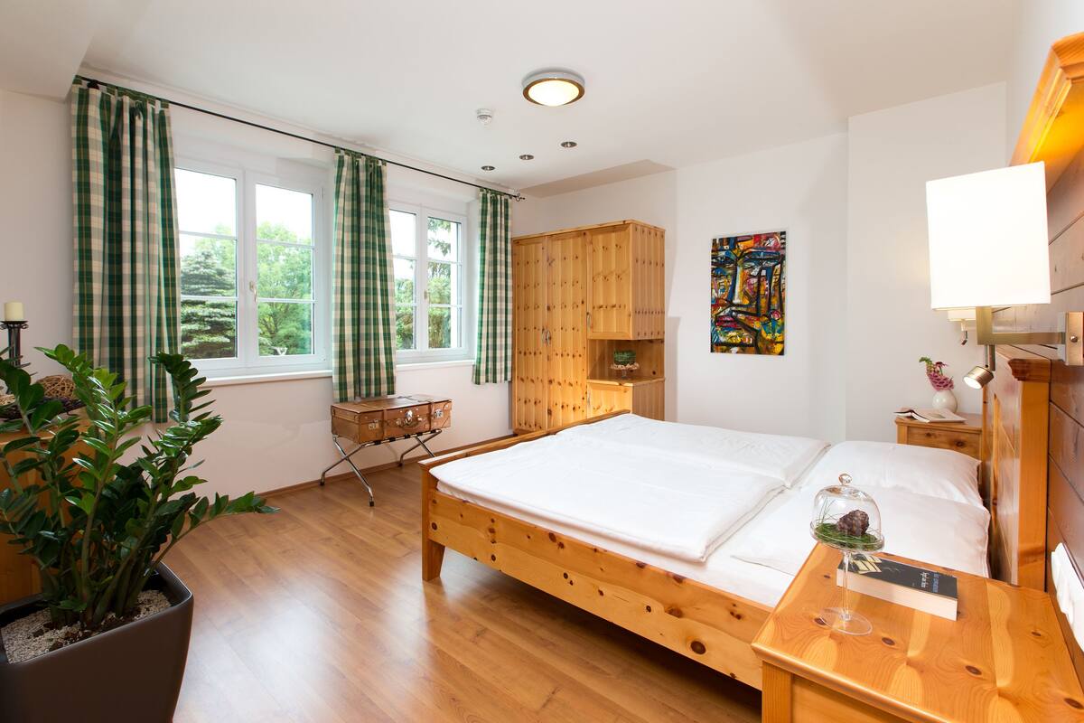 Un lit en bois avec du linge de lit blanc, une table de chevet avec une lampe, une armoire en bois, une fenêtre avec des rideaux et une plante en pot.