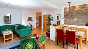 Ein modernes Wohnzimmer mit einem grünen Sofa, einem Holzespeisetisch und roten Stühlen. Es gibt eine kleine Küchenzeile mit einer Wandverkleidung aus Stein und einer Holztür.