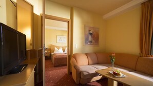 Habitación de hotel con televisor, sofá, una mesita con un florero y fruta, y una cama con almohada.