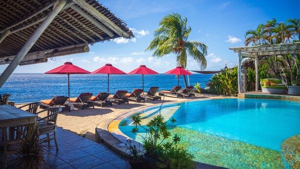 Un espace aménagé au bord de la piscine, avec des chaises longues et des parasols rouges, offrant une vue sur une mer d’un bleu limpide.
