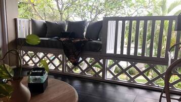 A wooden deck with a white lattice railing, a cushioned bench with pillows, a potted plant, and a small table with a vase and a box.