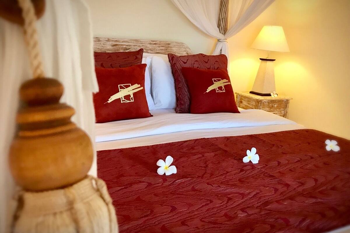 Een hotelkamer met een houten bedframe, rode en witte beddengoed, een bedlampje en een decoratief kussen met een gouden ontwerp.