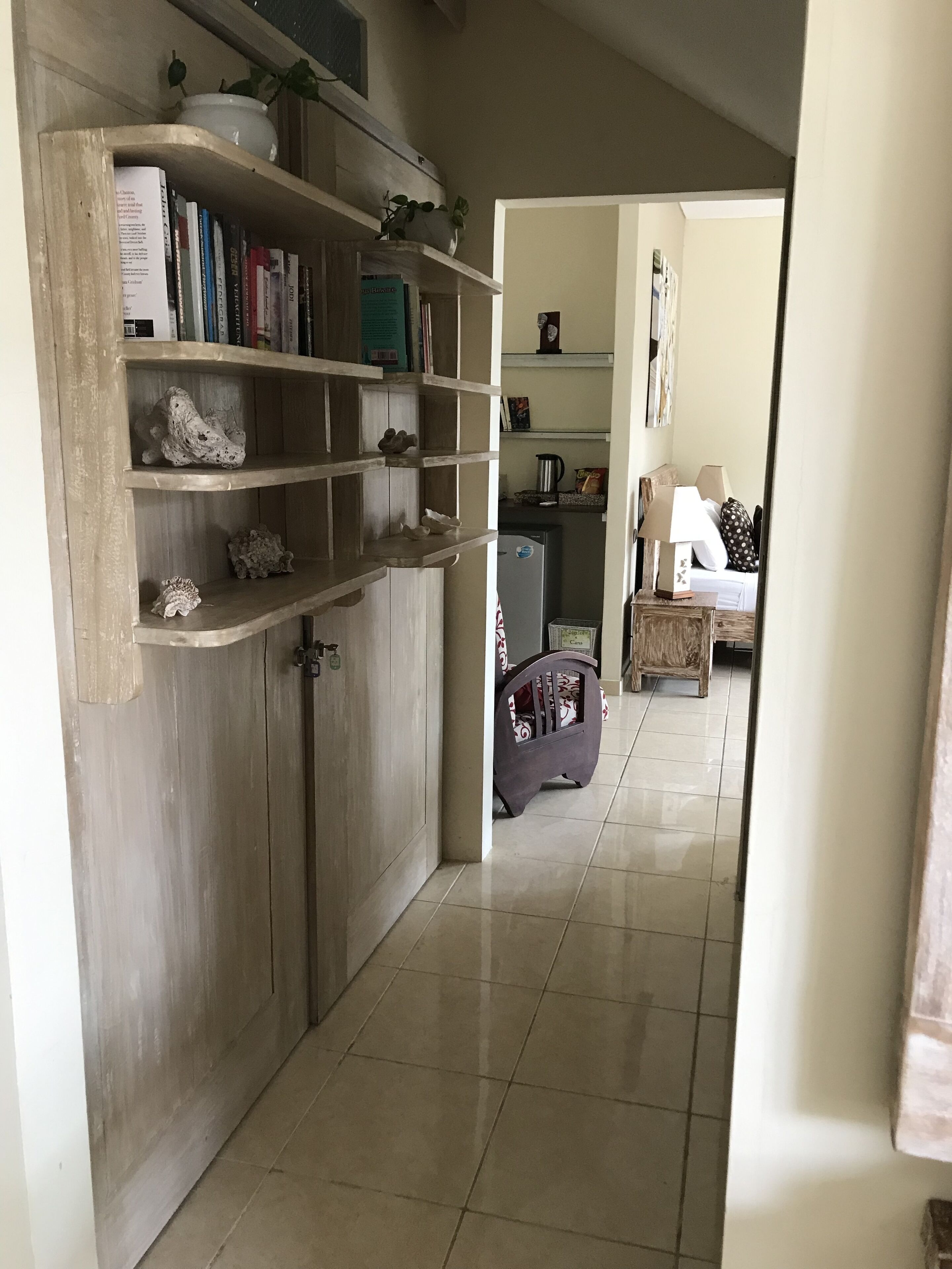 A hallway with a wooden wardrobe, shelves with books and decorative items, a kitchen area with a refrigerator, and a dining area with a table and chairs.