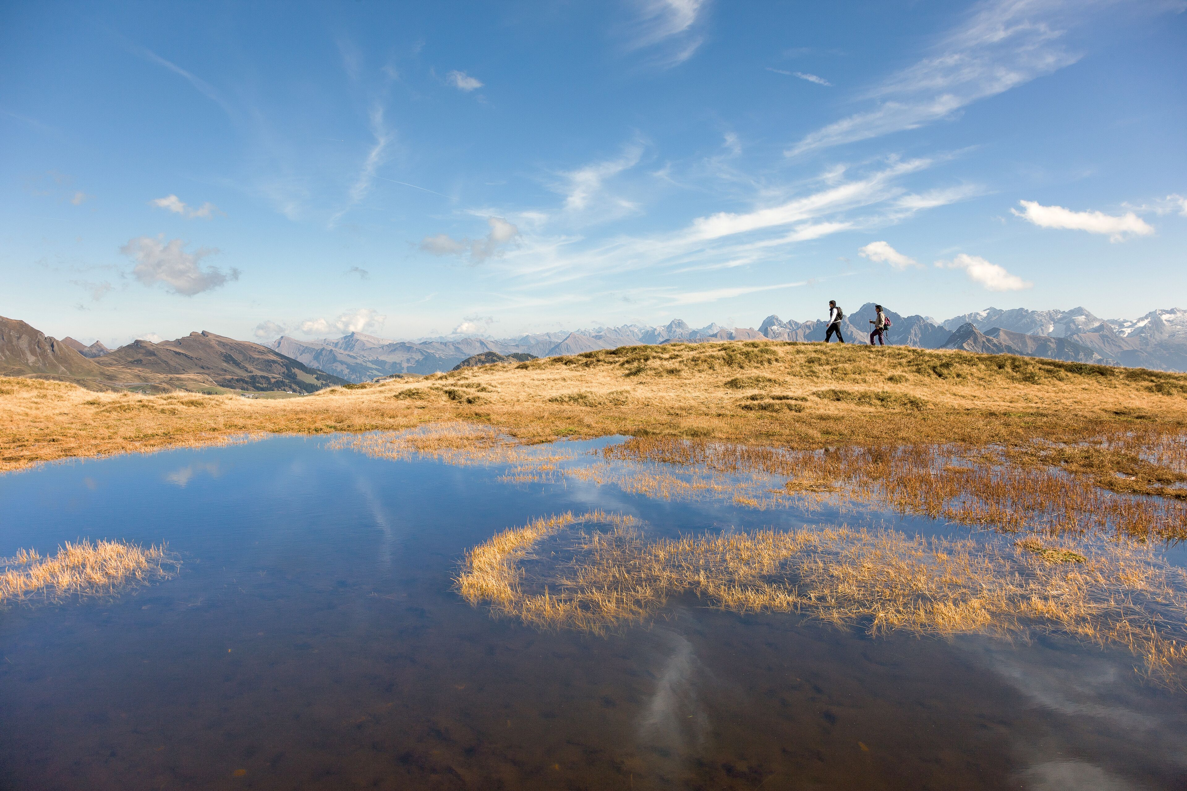 Ein weites Panorama einer Berglandschaft mit blauem Himmel und einem kleinen Gewässer, das die umliegende Landschaft spiegelt.