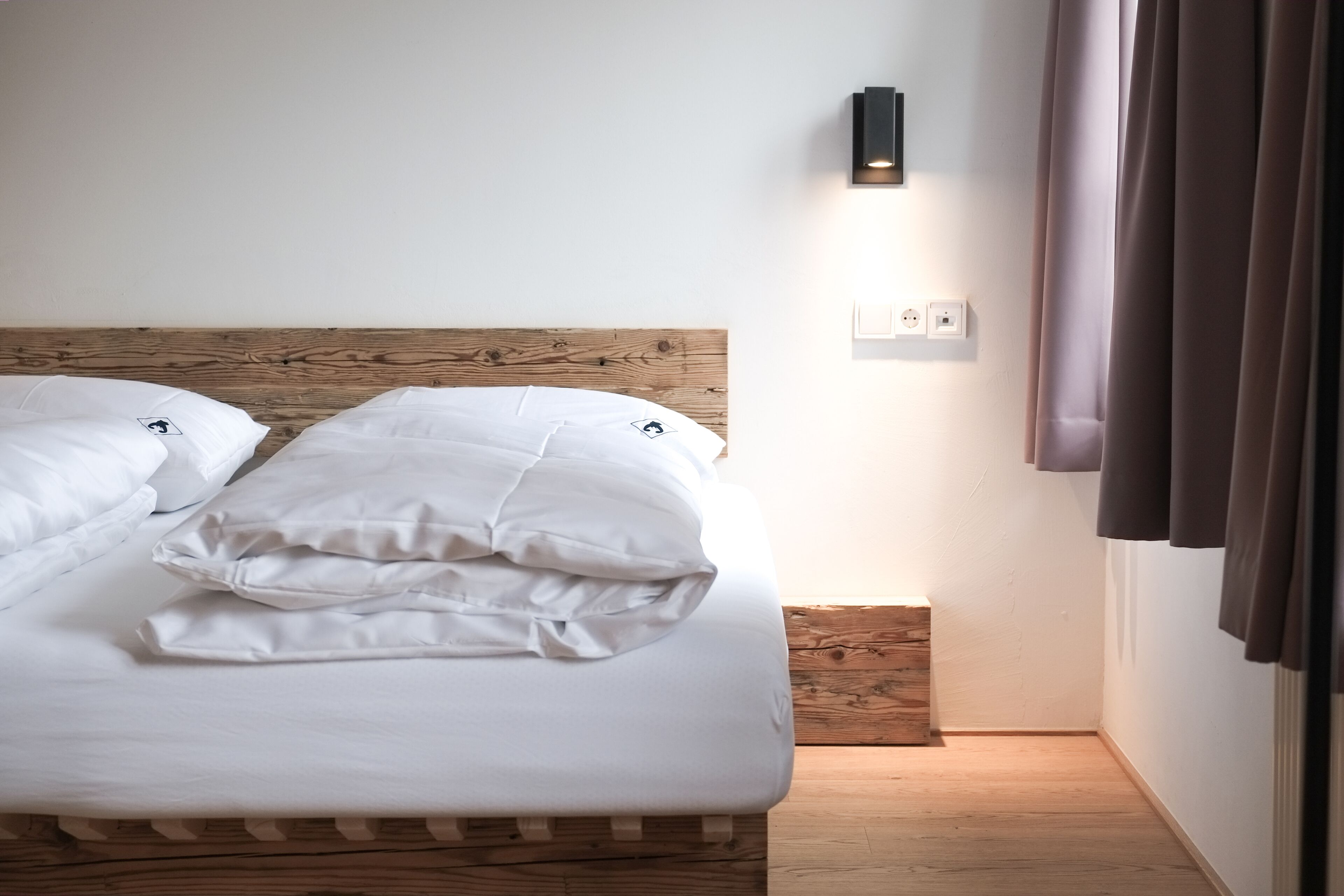 Una cama bien tendida con sábanas blancas, un cabecero de madera, un aplique en la pared y un enchufe.