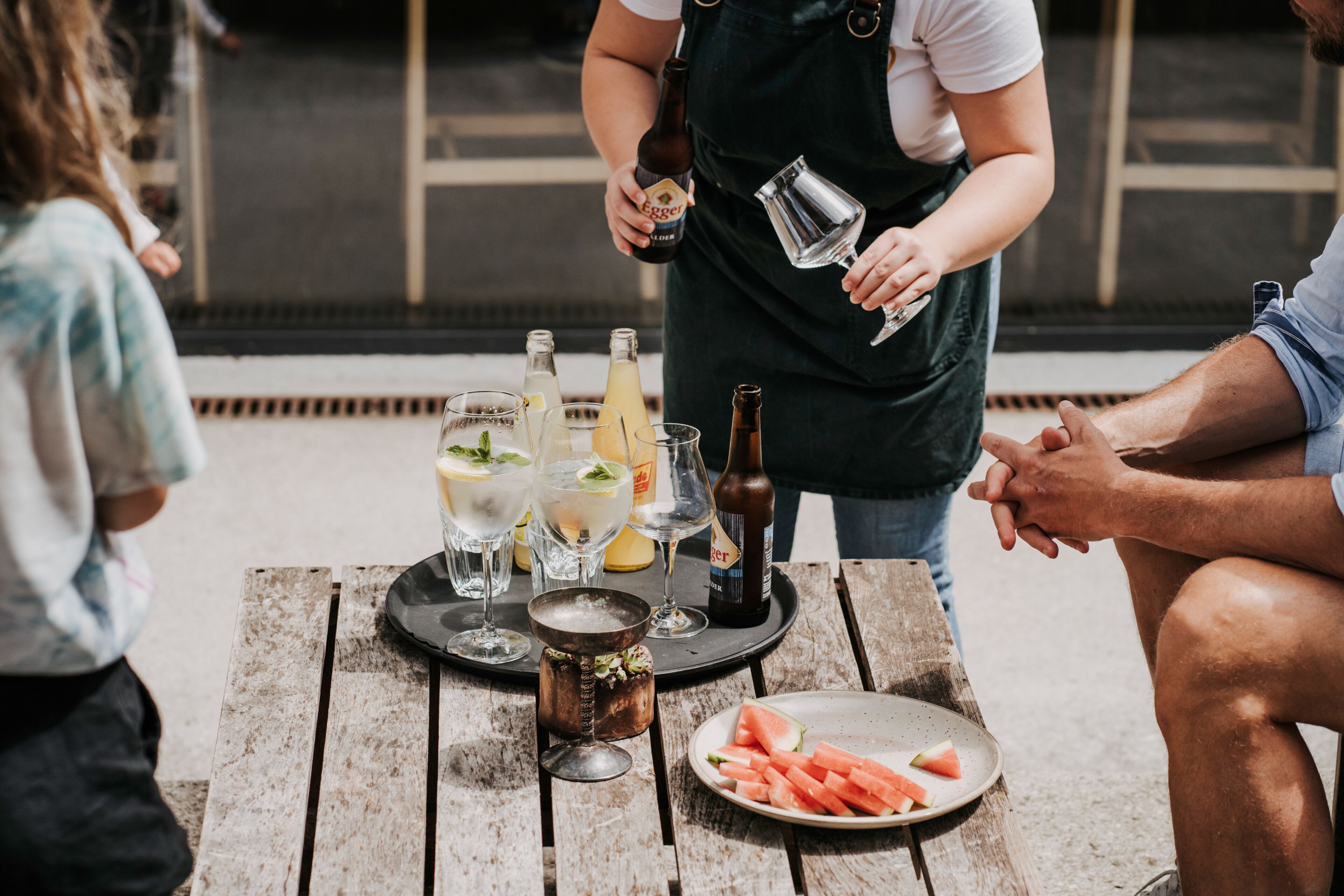 A wooden table with a variety of drinks and a watermelon slice, indicating a casual outdoor dining setting.
