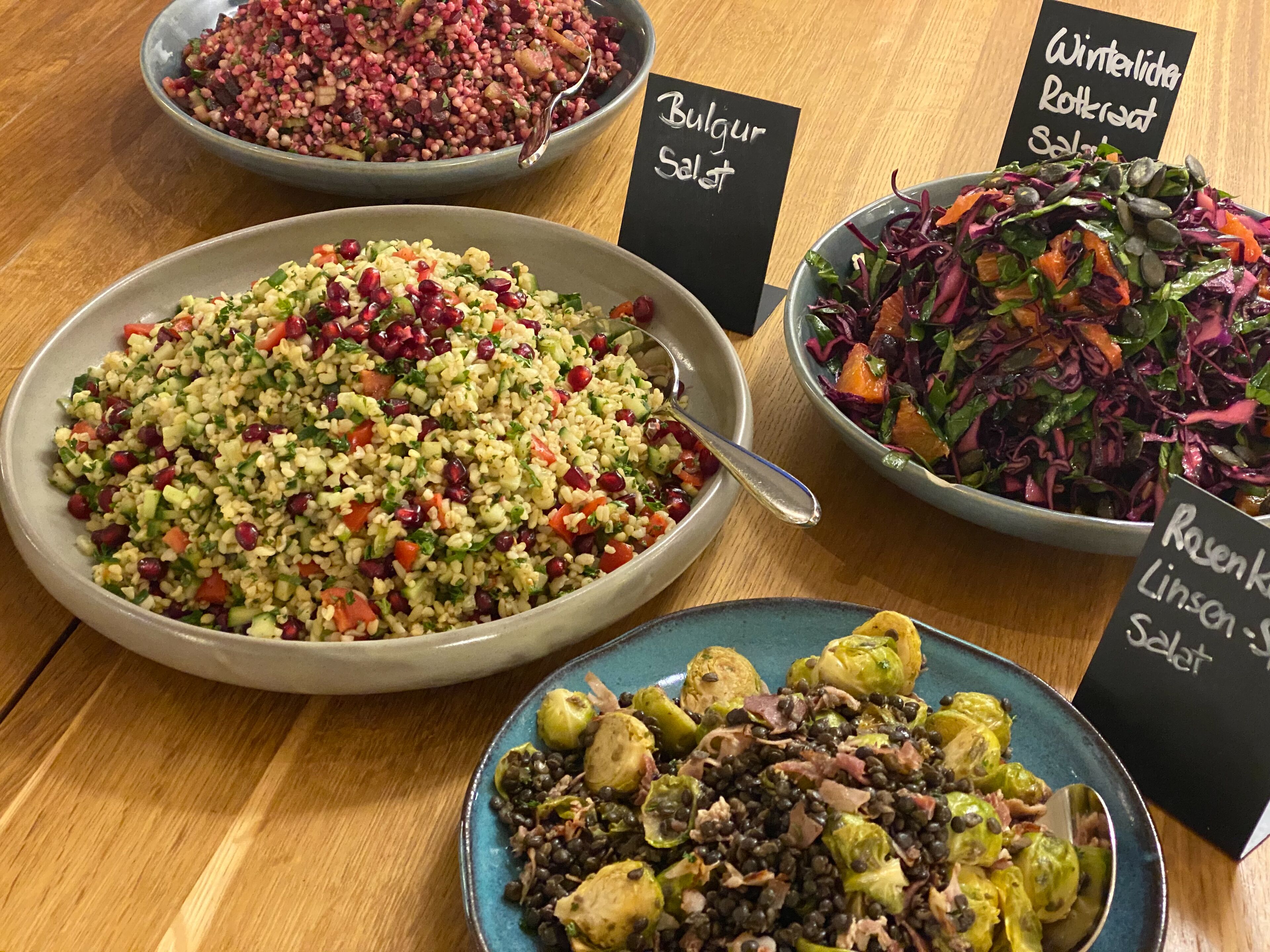 A table with various dishes labeled in German, including 'Bulgur Salad' and 'Ravenkuchen Linsen-Salat'.