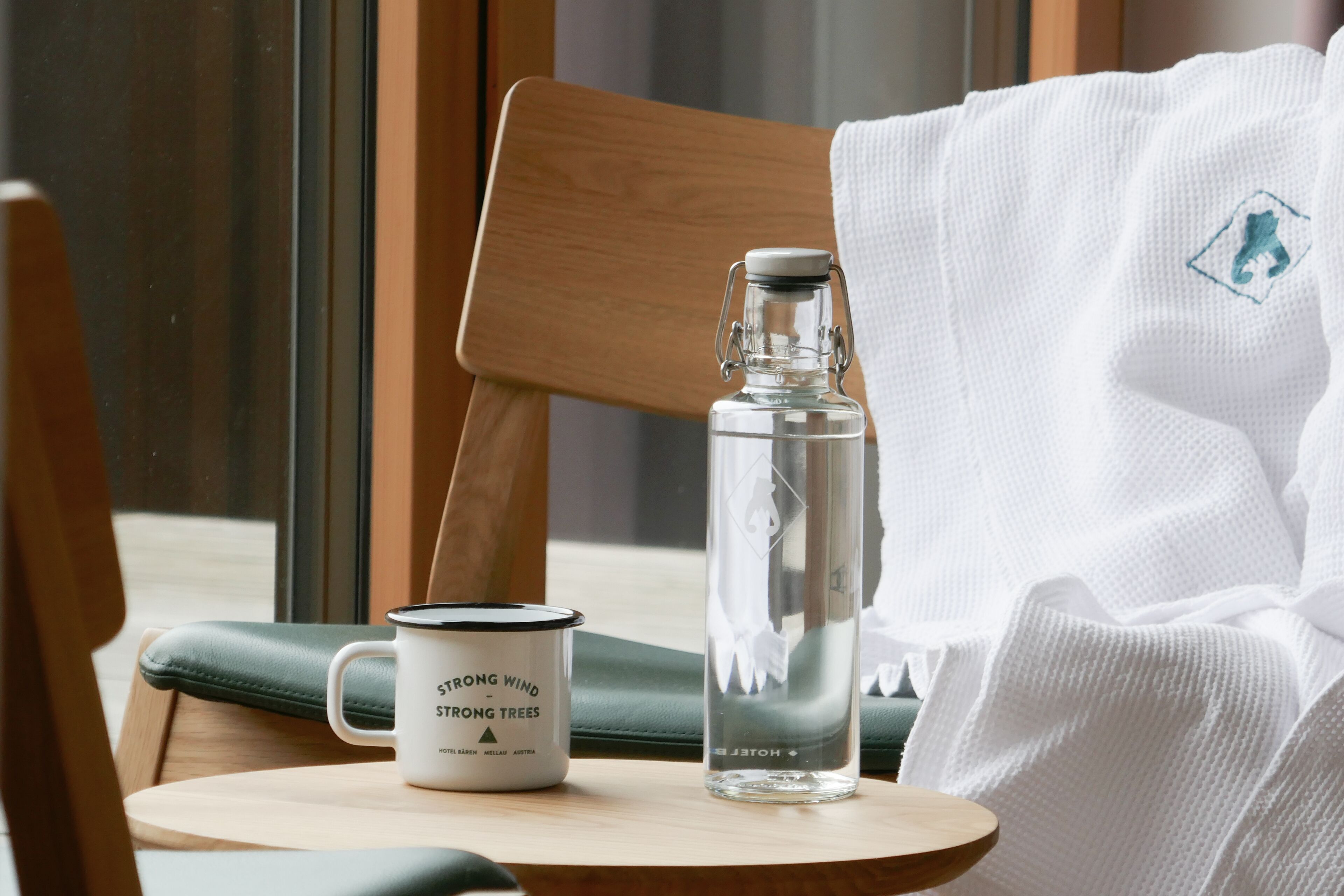 Eine klare Glasflasche, eine weiße Tasse und ein Holzstuhl in einem Raum mit Fenster und weißer Decke.