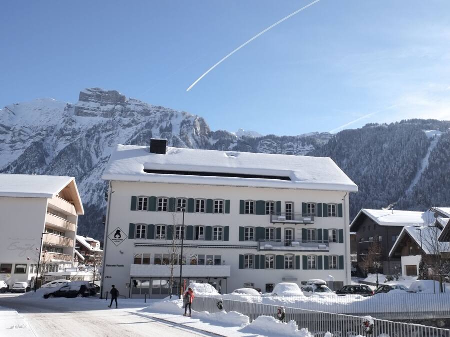Un edificio de varios pisos con techo nevado y un complejo de esquí al fondo.