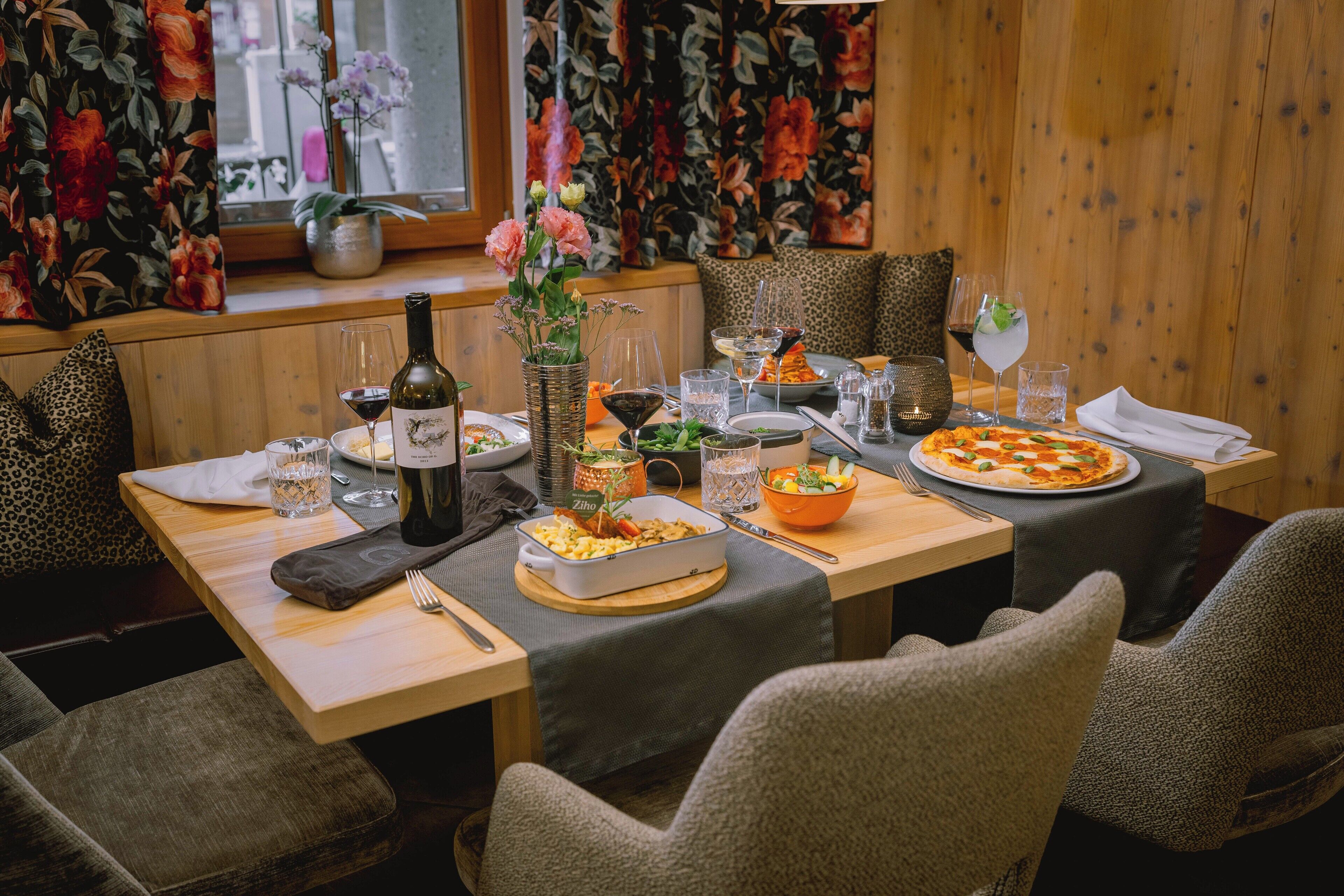 Une table à manger dressée avec des assiettes, des verres et une bouteille de vin, entourée de chaises et d’une fenêtre ornée de rideaux à motifs floraux.