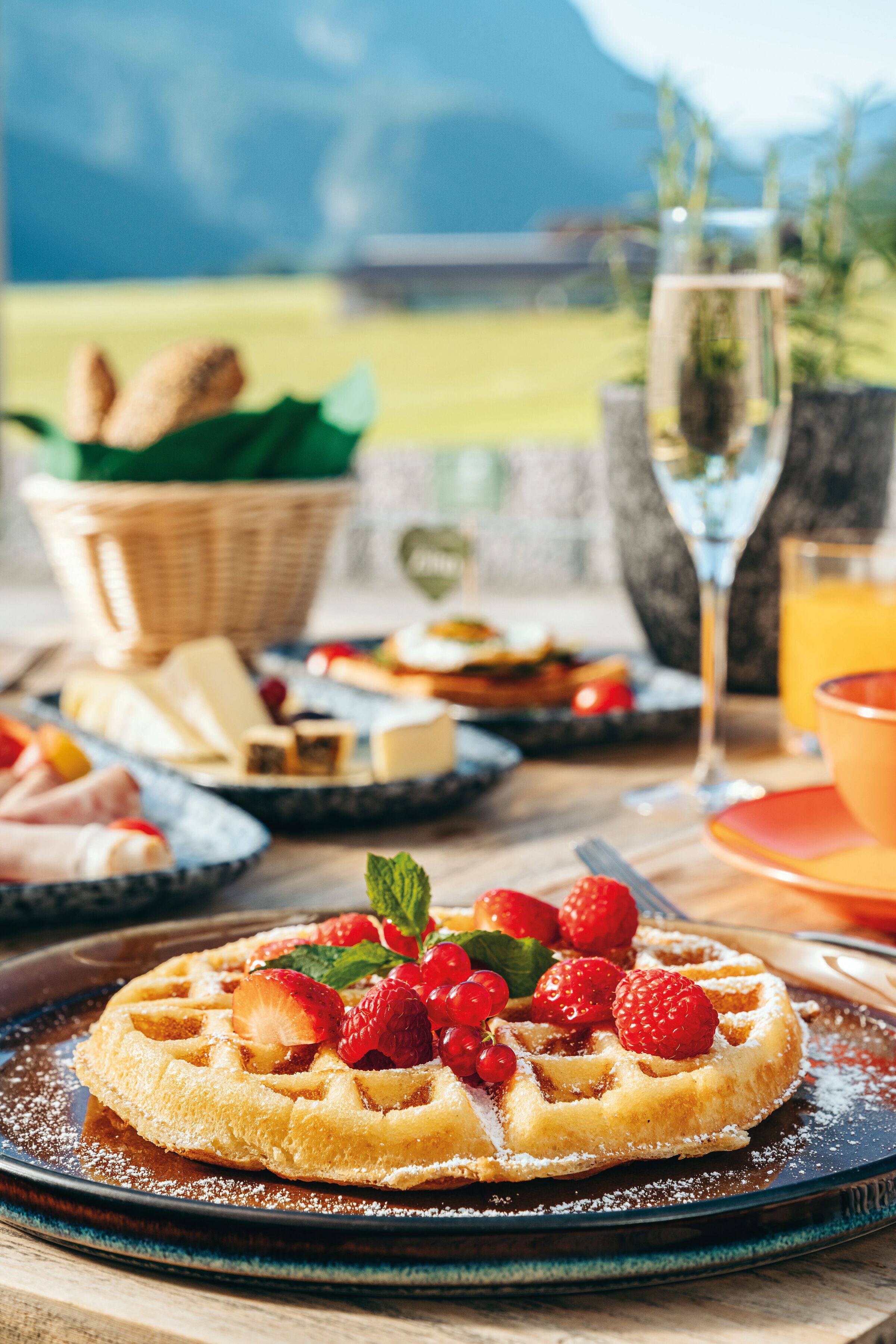 A breakfast setting with waffles topped with strawberries and mint, a glass of orange juice, and a basket of bread and cheese.