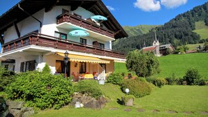 Un chalet à deux étages doté d’un balcon, d’un coin repas avec des sièges extérieurs, et offrant une vue sur une église et des montagnes en arrière-plan.