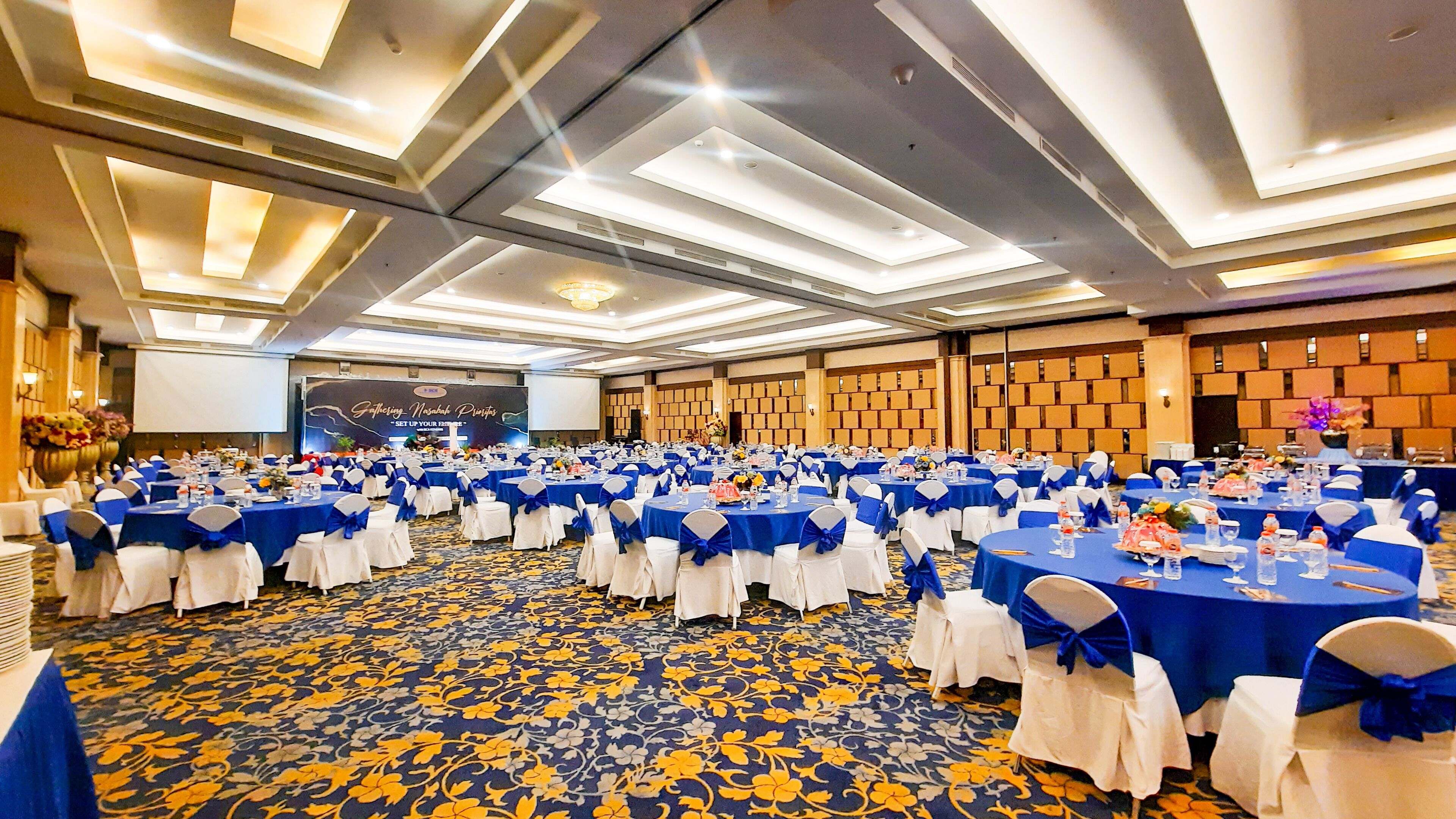 A banquet hall with round tables covered in blue tablecloths and white chairs, a floral carpet, and a stage with a presentation screen.