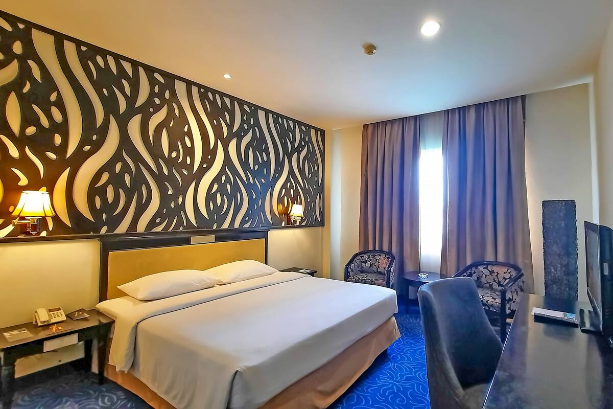 A hotel room with a large bed, two chairs, a desk with a lamp, a TV, and a decorative wall panel.