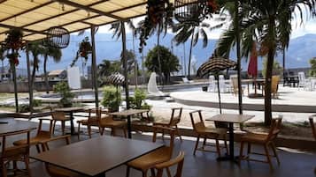 An outdoor seating area with tables and chairs, a covered section with a roof, and a view of the ocean and mountains in the background.