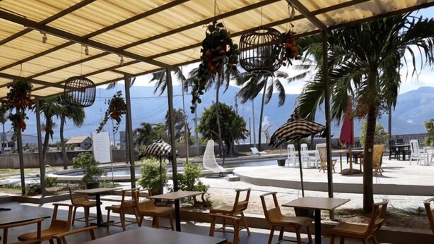 An outdoor seating area with tables and chairs, a covered section with a roof, and a view of the ocean and mountains in the background. - Palu Golden (Palu)