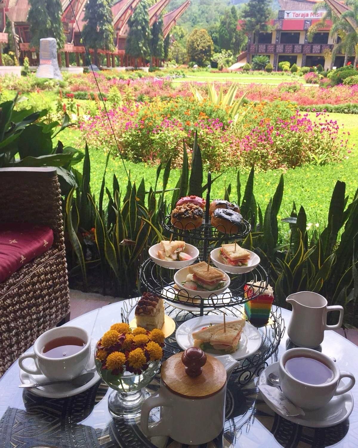 A table set with a variety of pastries, sandwiches, and a cake, with a garden view in the background.