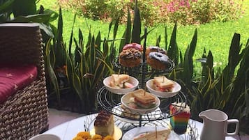 A table set with a variety of pastries, sandwiches, and a cake, with a garden view in the background.