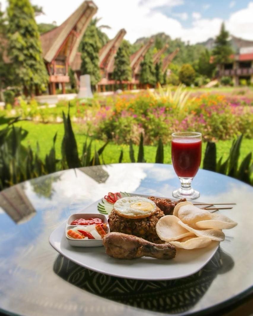 A table with a meal consisting of meat, eggs, chips, and a drink, set against a backdrop of a garden with trees and a building.