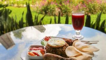 A table with a meal consisting of meat, eggs, chips, and a drink, set against a backdrop of a garden with trees and a building.