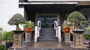 A building entrance with a staircase leading up to a glass door, flanked by potted bonsai trees on pedestals. - Amalia Lampung (Bandar Lampung)