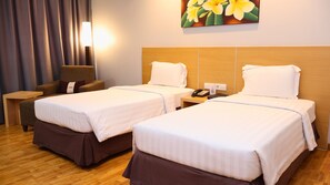 A hotel room with two beds, a wooden headboard, a nightstand with a phone and a lamp, a chair, and a painting of flowers on the wall.