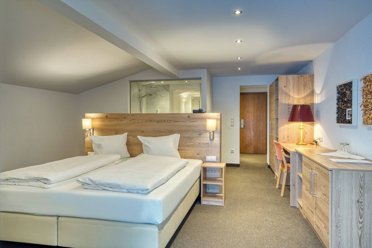 A modern hotel room with a large bed, a wooden headboard, a desk with a chair, and a vanity with a mirror.