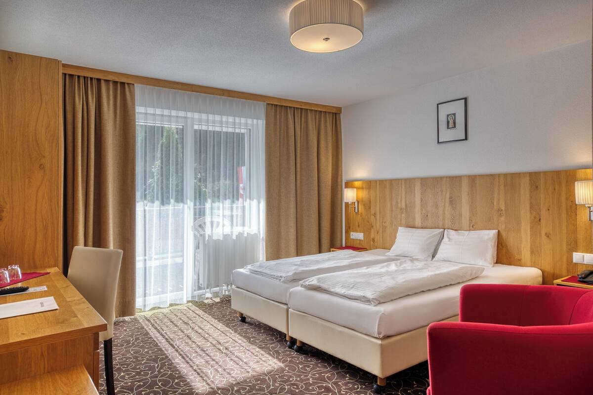 A hotel room with a wooden headboard, two beds, a desk with a chair, and a red armchair.