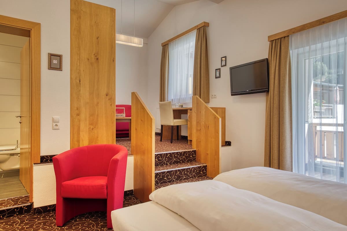 A hotel room with a bed, a red chair, a desk with a chair, a TV mounted on the wall, and a staircase leading to an upper level.