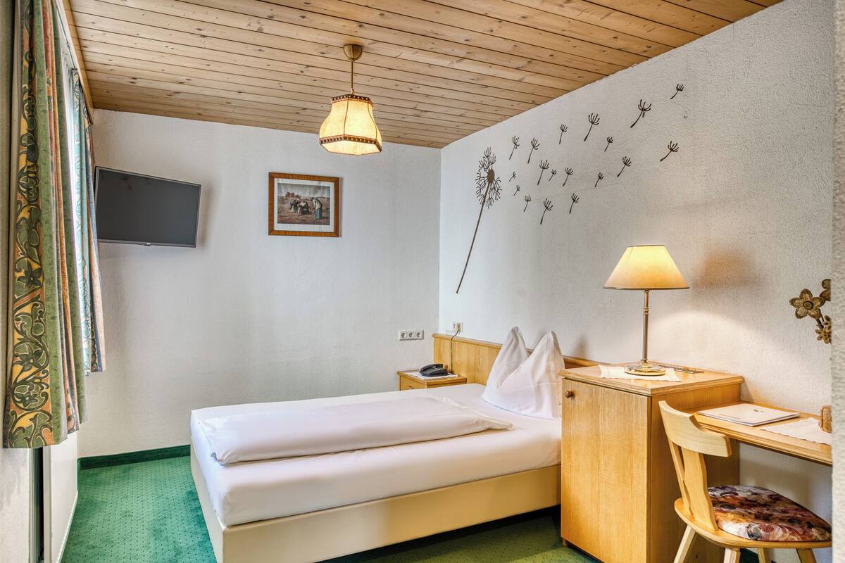A hotel room with a bed, a desk, a chair, a lamp, a TV, and a wall decoration.