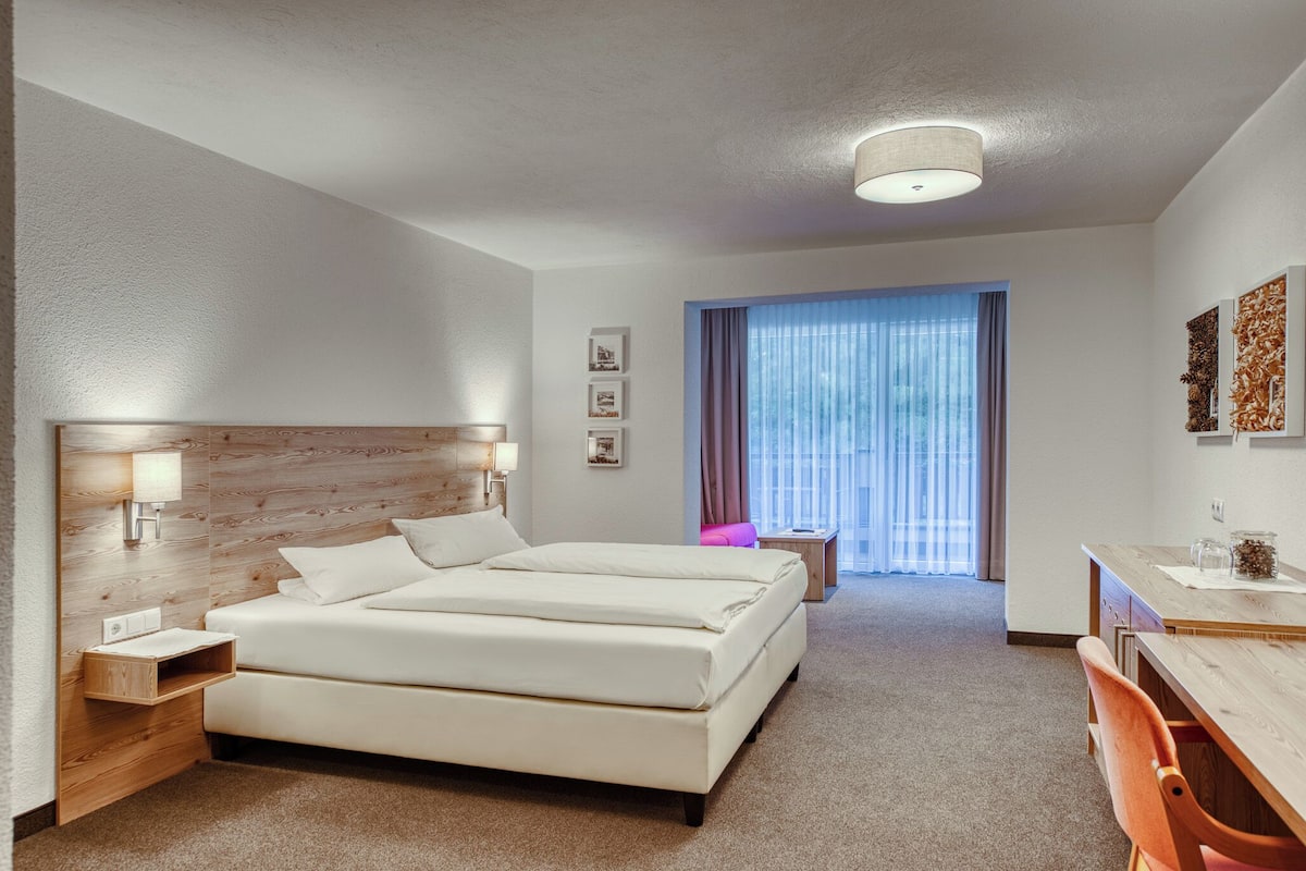 A hotel room with a large bed, bedside tables, a desk, and a chair. There is a window with curtains and a view of the outside.