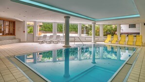 An indoor swimming pool with clear blue water, surrounded by white and yellow lounge chairs, and large windows offering views of greenery outside. - Hotel Erika (Nauders)