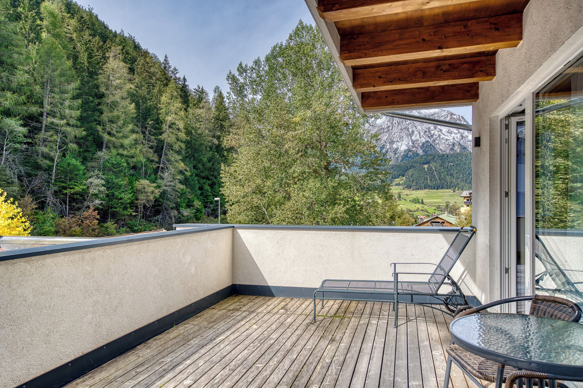 Un balcón con cubierta de madera, una mesa de vidrio y sillas, con vista a un área boscosa y montañas.