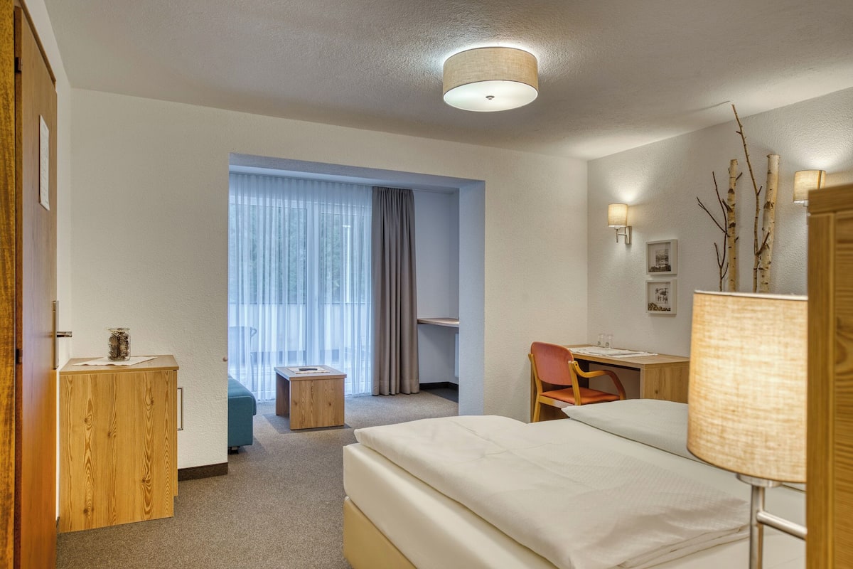 A hotel room with a bed, a desk, a chair, a small table, and a lamp.