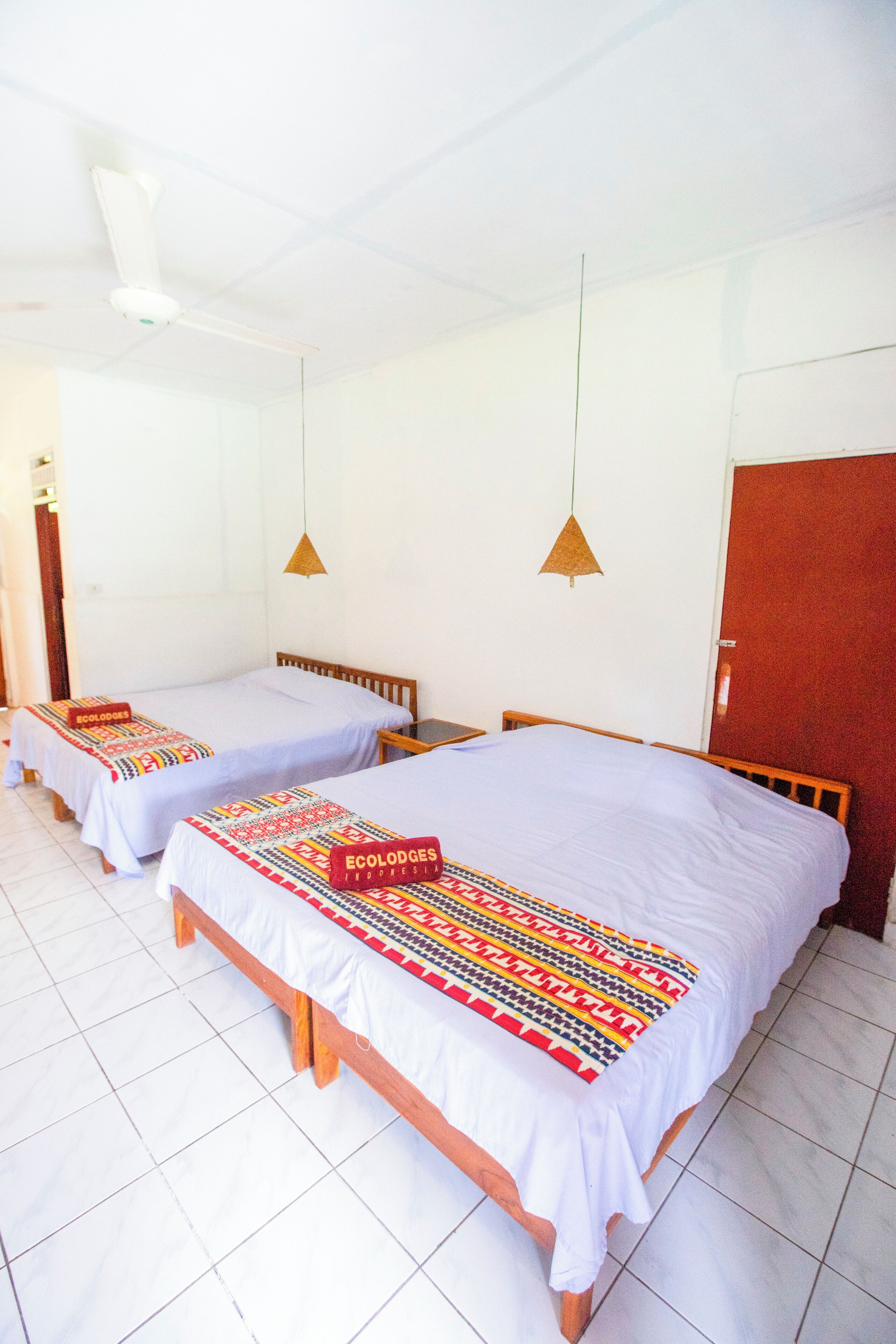 A hotel room with two single beds, each with a white bedspread and a colorful blanket. There are two pendant lights hanging from the ceiling, and a wooden door on the right.
