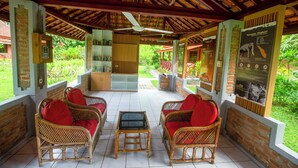 A covered outdoor seating area with wicker furniture and red cushions, a ceiling fan, and informational posters on the walls. - The Satwa Elephant Ecolodge (Labuhan Ratu)