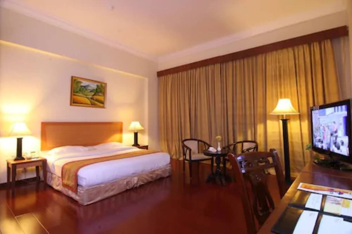 A hotel room with a bed, two bedside lamps, a television, a desk with a chair, and a large window with curtains.