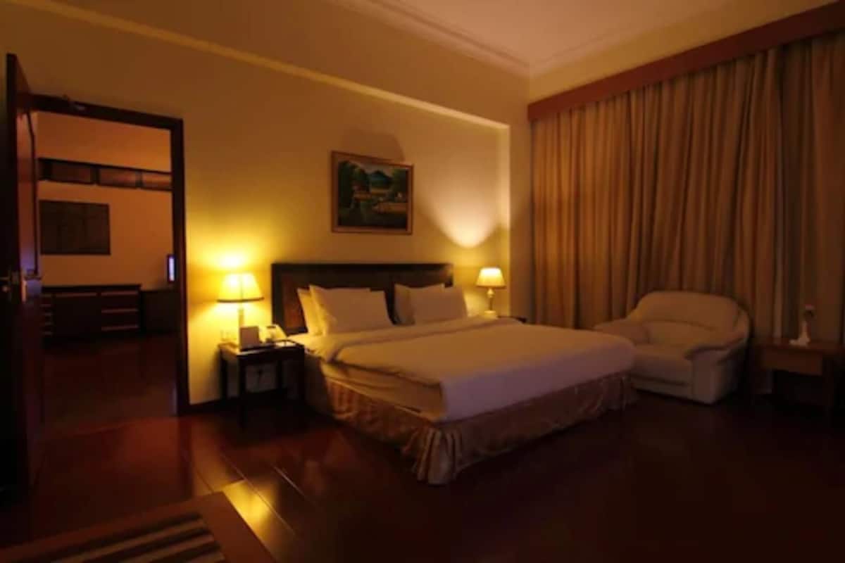 A hotel room with a large bed, two bedside tables with lamps, a chair, a painting on the wall, and a door leading to another room.